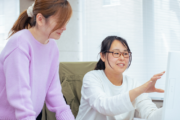 2人の女性がパソコンを使って作業をしており、1人が画面を指さしながら説明をしています。